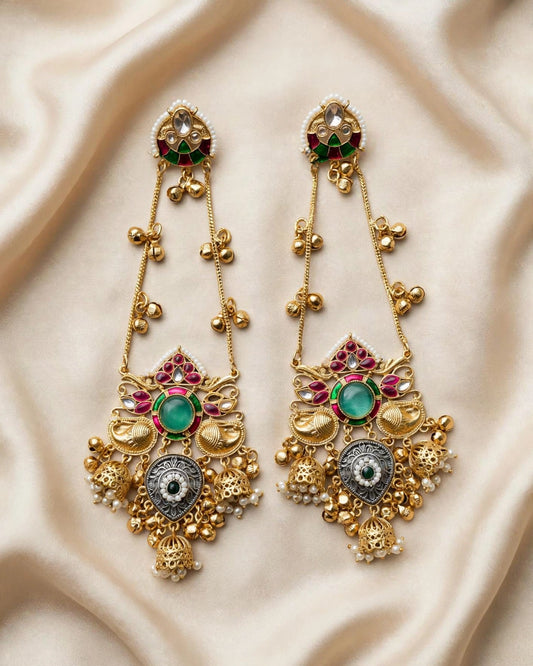 A pair of ornate gold-toned traditional Indian chandelier earrings with green and red stones, set against a draped beige satin fabric background.