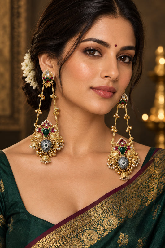 A pair of ornate gold-toned Indian chandbali earrings with green and red enamel details, set against a draped cream satin fabric background.